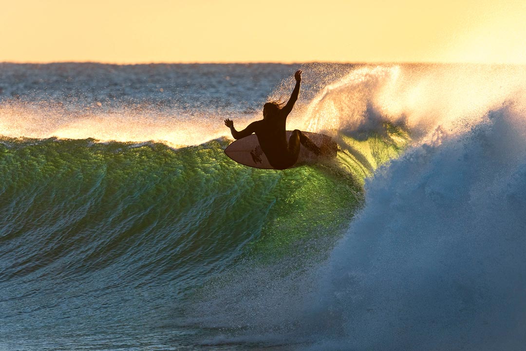 A surfer throws spray off the lip of a back-lit wave.
