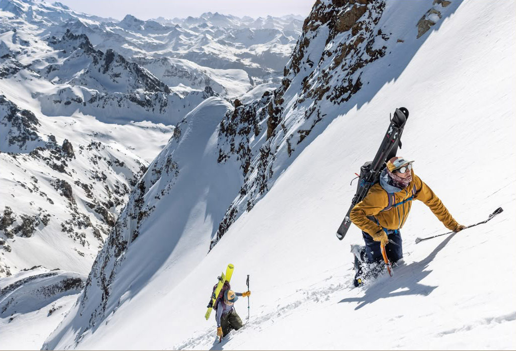 Two people climb a steep, snowy mountain with skis on their backs.
