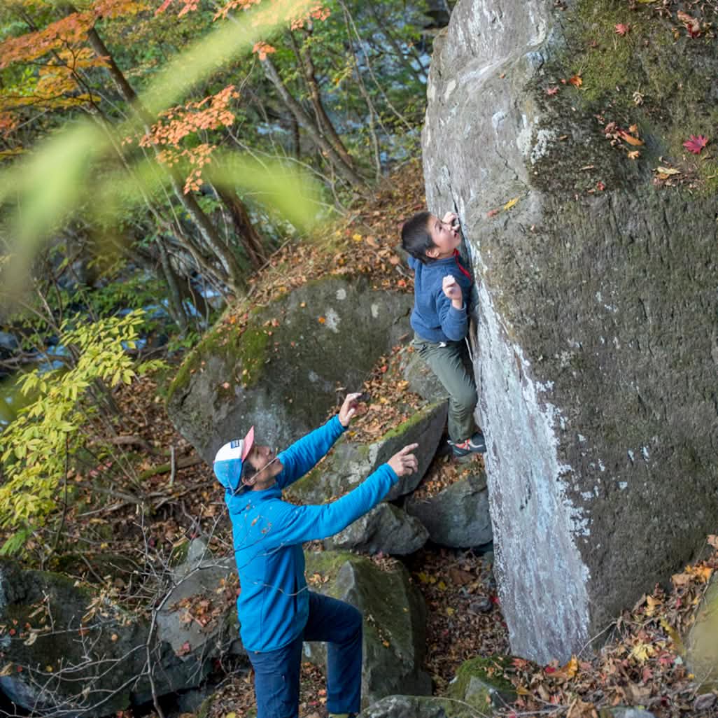 A kid climbs a boulder while his dad provides a spot.