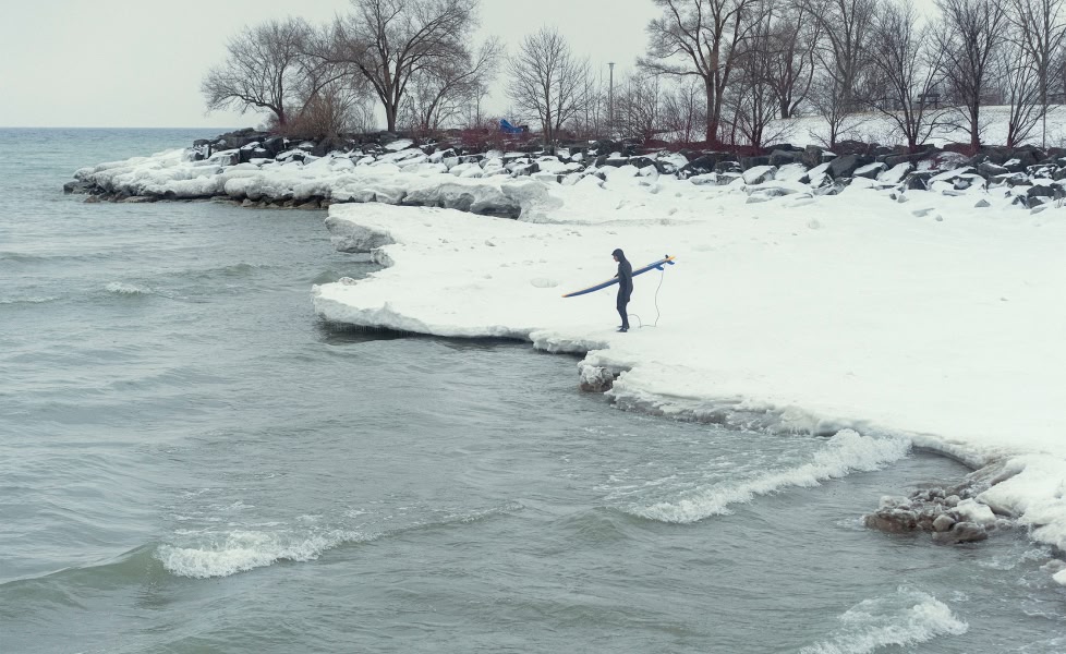 A surfer stands in the snow preparing to paddle out in a cold lake.