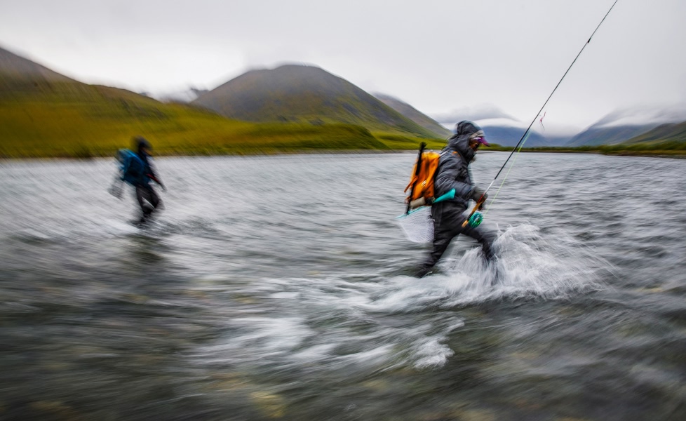 A blurry photo of two anglers wading a wide river with their fly rods and gear.
