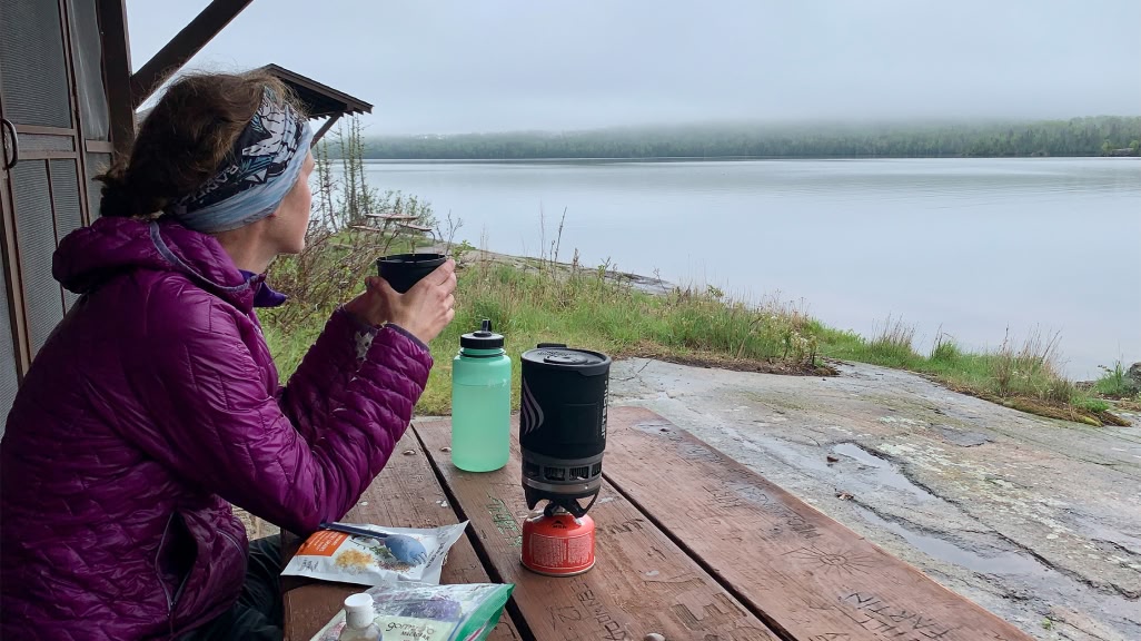 A person sits at a picnic table and looks at a misty lake while drinking tea.