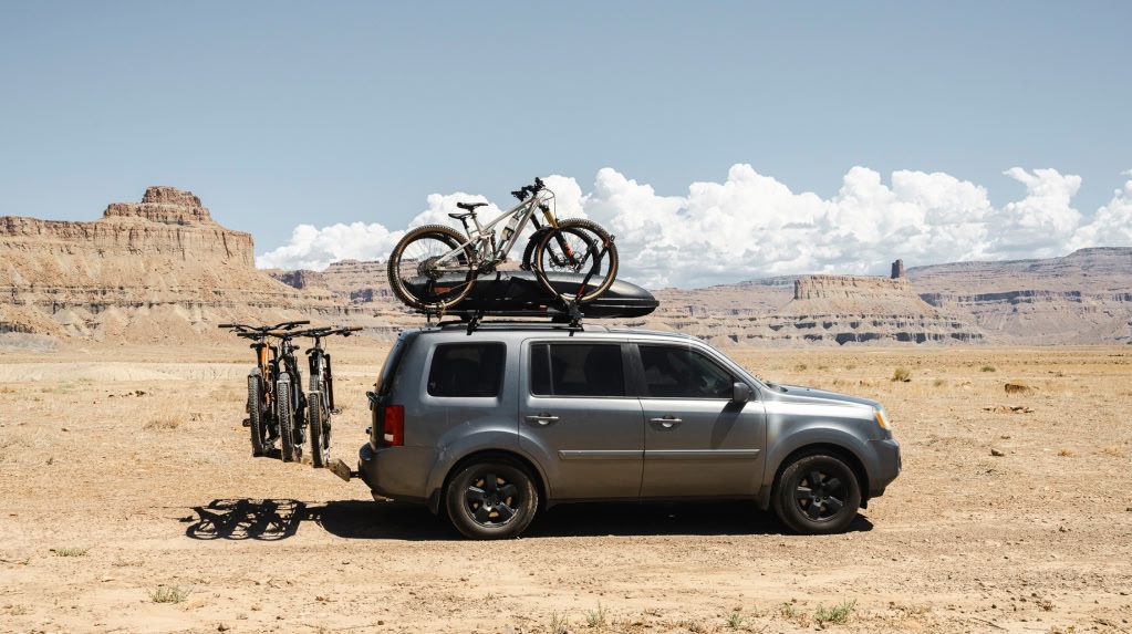 A silver car, loaded up with bikes on the roof and bikes on the back rack, is parked in the desert.