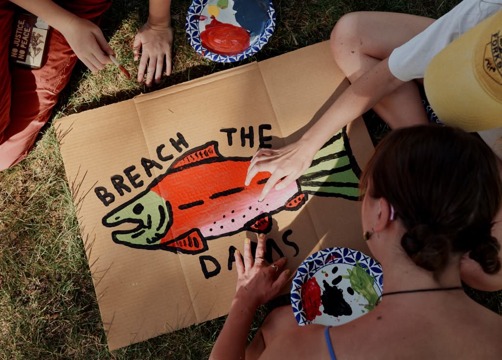 Patagonia Action Works. Keep Showing Up. Because Earth has always shown up for us. Three people sit on grass and paint a protest sign with a fish and the statement “Breach the Dams.”