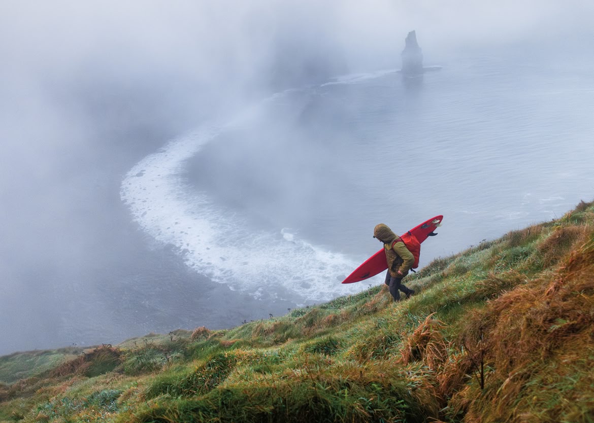 A person holding a surfboard hikes down a steep trail to a foggy coastline.
