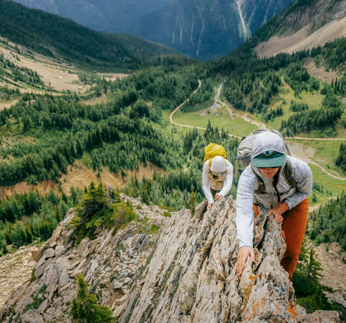 Two people hike up a rocky spine high above a pine forest valley.