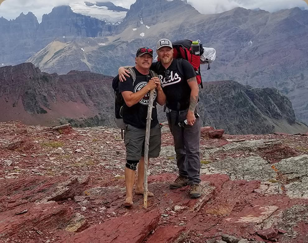 Two people wearing backpacks stand on a red rock mountain ridge.