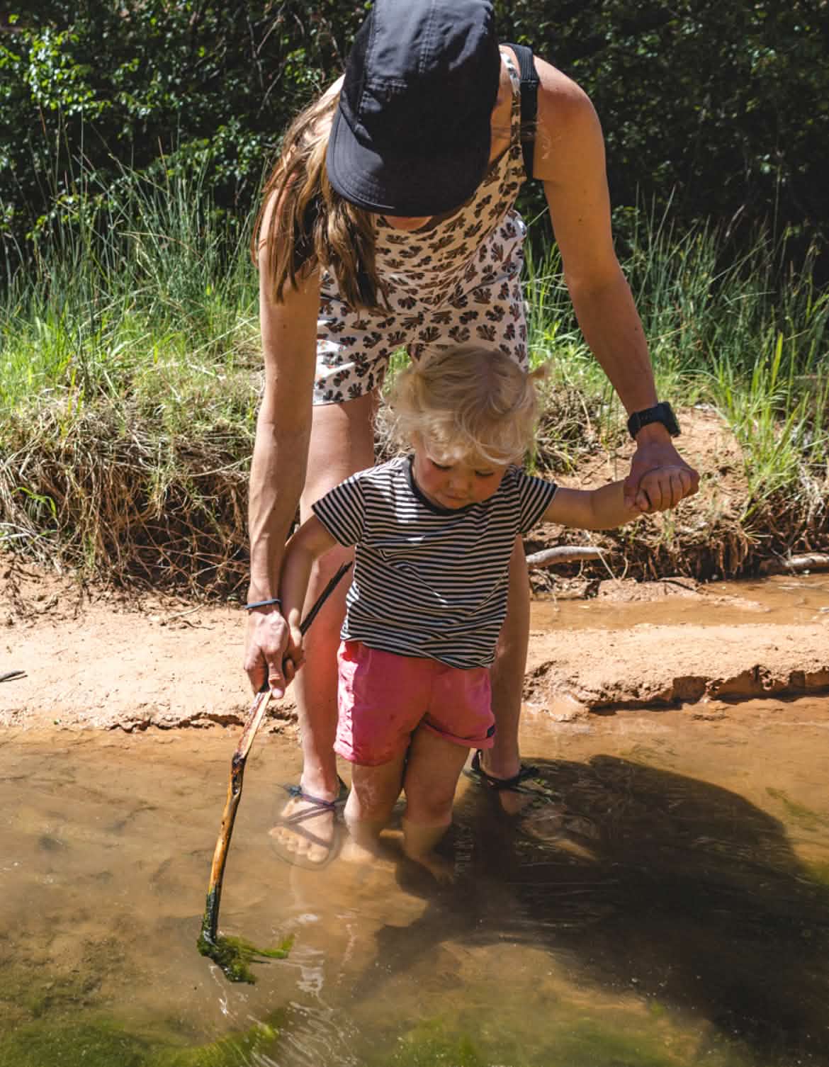 Patagonia. A person helps a young person balance as they stand in a creek and poke at algae with a stick.