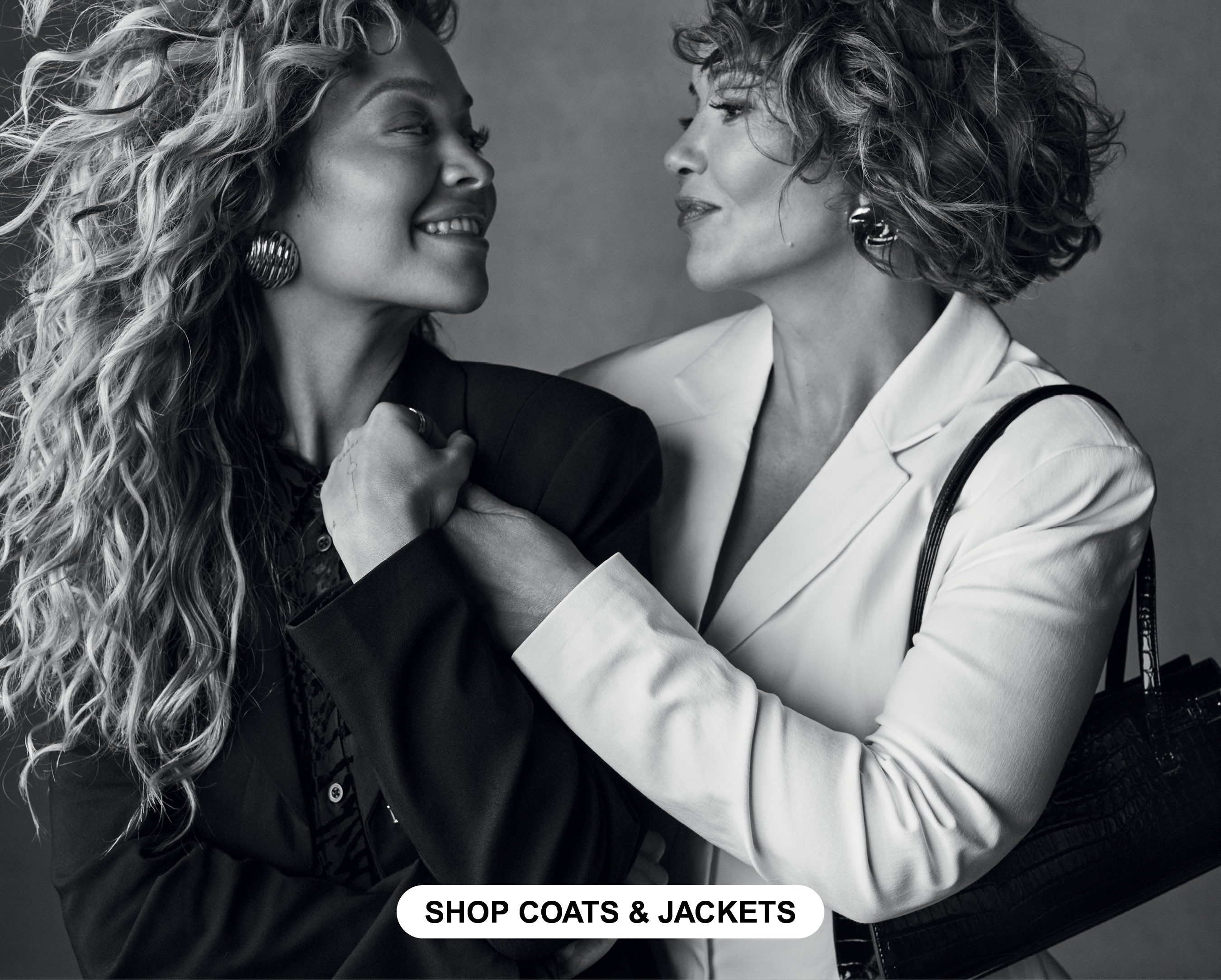 Rita Ora and Vera wearing a black blazer and a white blazer smiling at each other. 