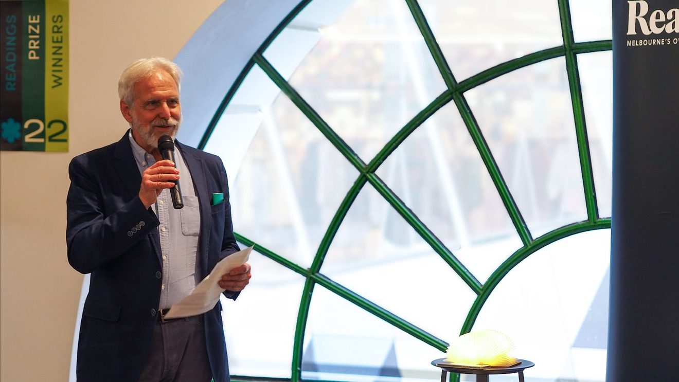 Mark Rubbo with a microphone standing in front of the window in our Emporium shop, announcing the 2022 Readings Prize winners.