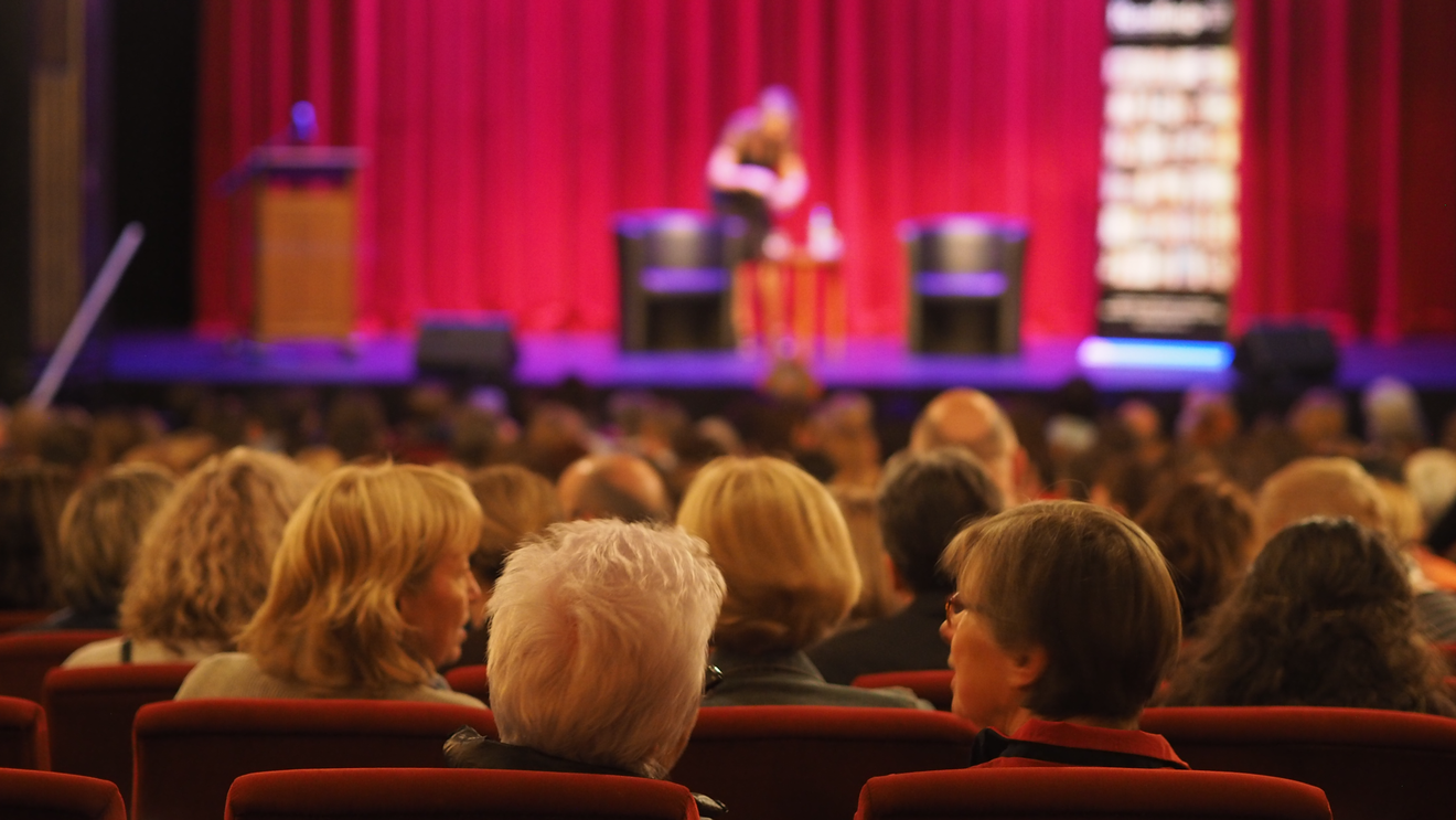 The backs of a seated audience, looking towards a theatre stage.