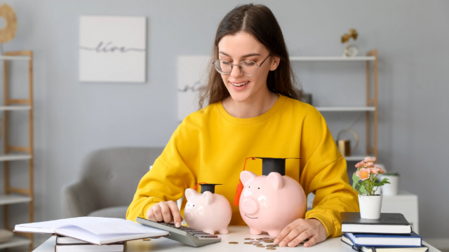 Student wearing a yellow sweatshirt sitting at a desk with 2 piggy banks, a calculator, books and coins on it. 