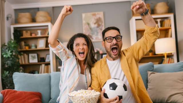 Couple sitting on a a couch, popcorn bowl in hand of woman, man is holding a soccer ball. They have their arms in the air cheering. 