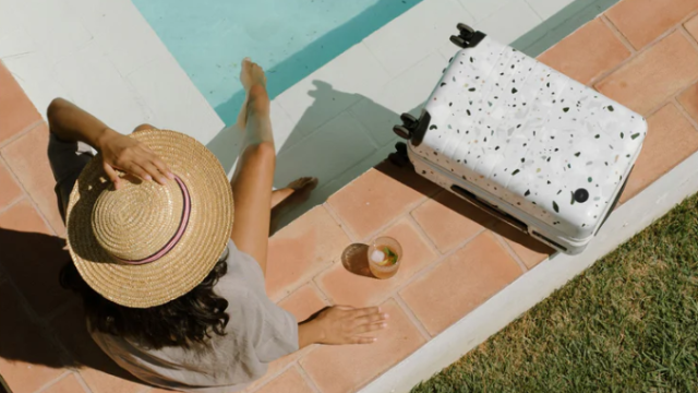 Woman sitting on the edge of a pool with a drink to her left and a monos suitcase beside that. 