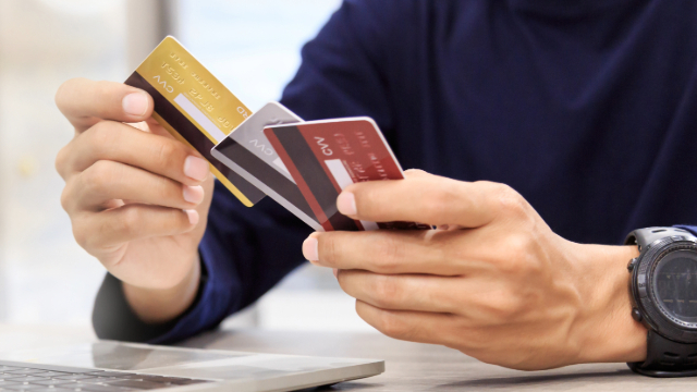 Person holding three credit cards in their hands. There is a laptop on the table and they are wearing a blue long sleeve shirt and a chunky black watch on their left wrist. 