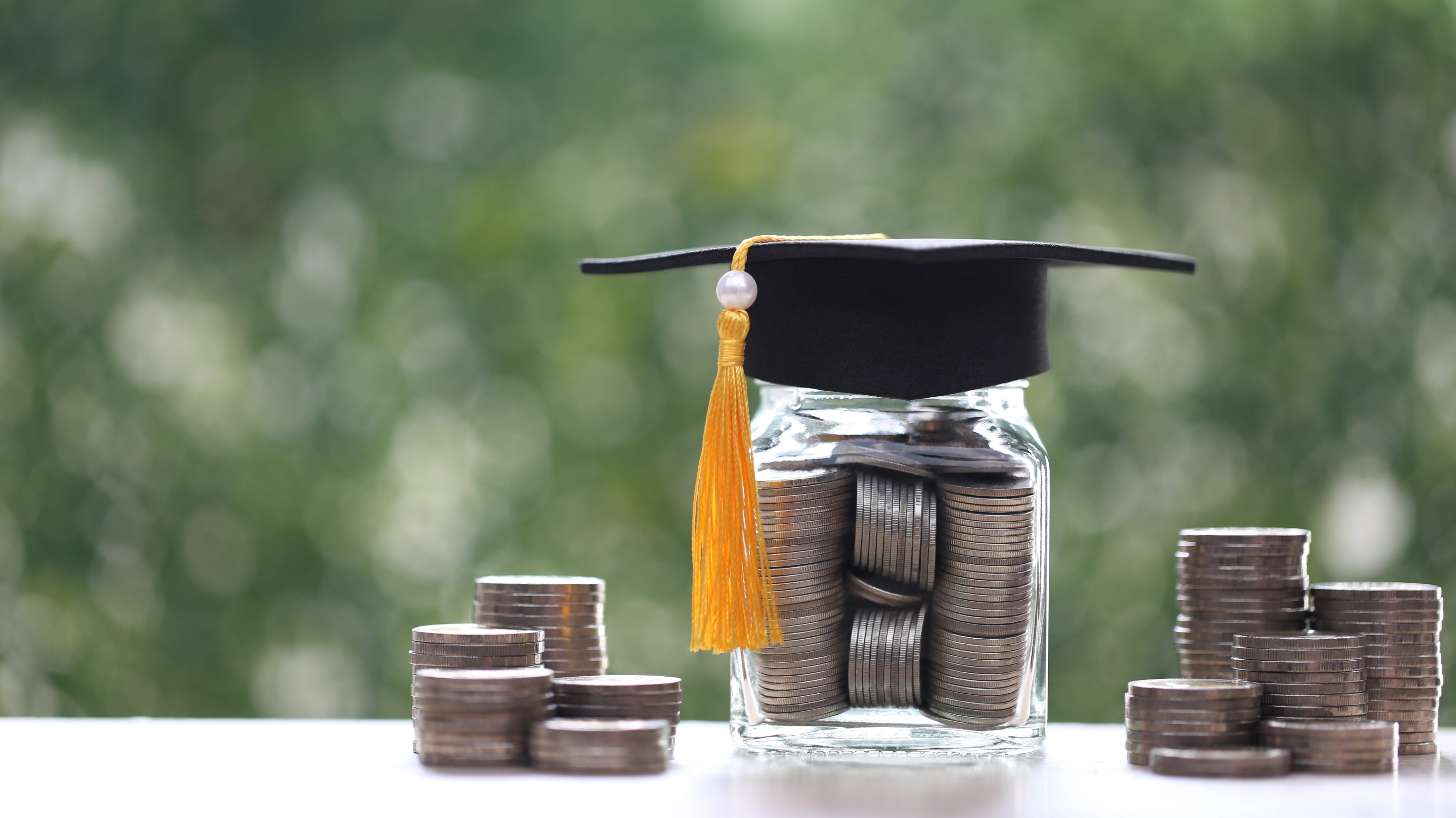 A jar filled with silver coins and a grad cap on top (yellow tassel) sits on a counter top or table. There are stacks of silver coins on either side and a blurred green background. 