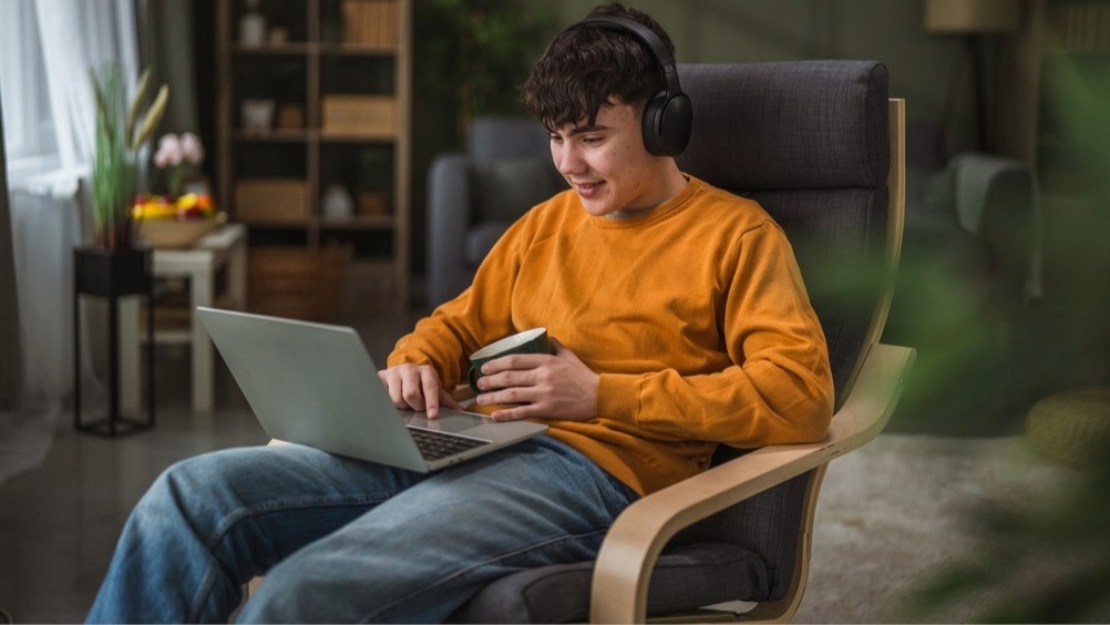 Young man sitting in a Poang chair from IKEA. He is wearing an orange sweatshirt, over the ear headphones and he has his laptop open on his lap, holding a black coffee mug in his left hand. 