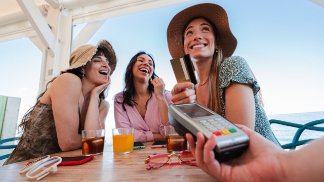 THreee young women on a beach patio having drinks. One of them is paying with her credit card on a portable debit machine. 
