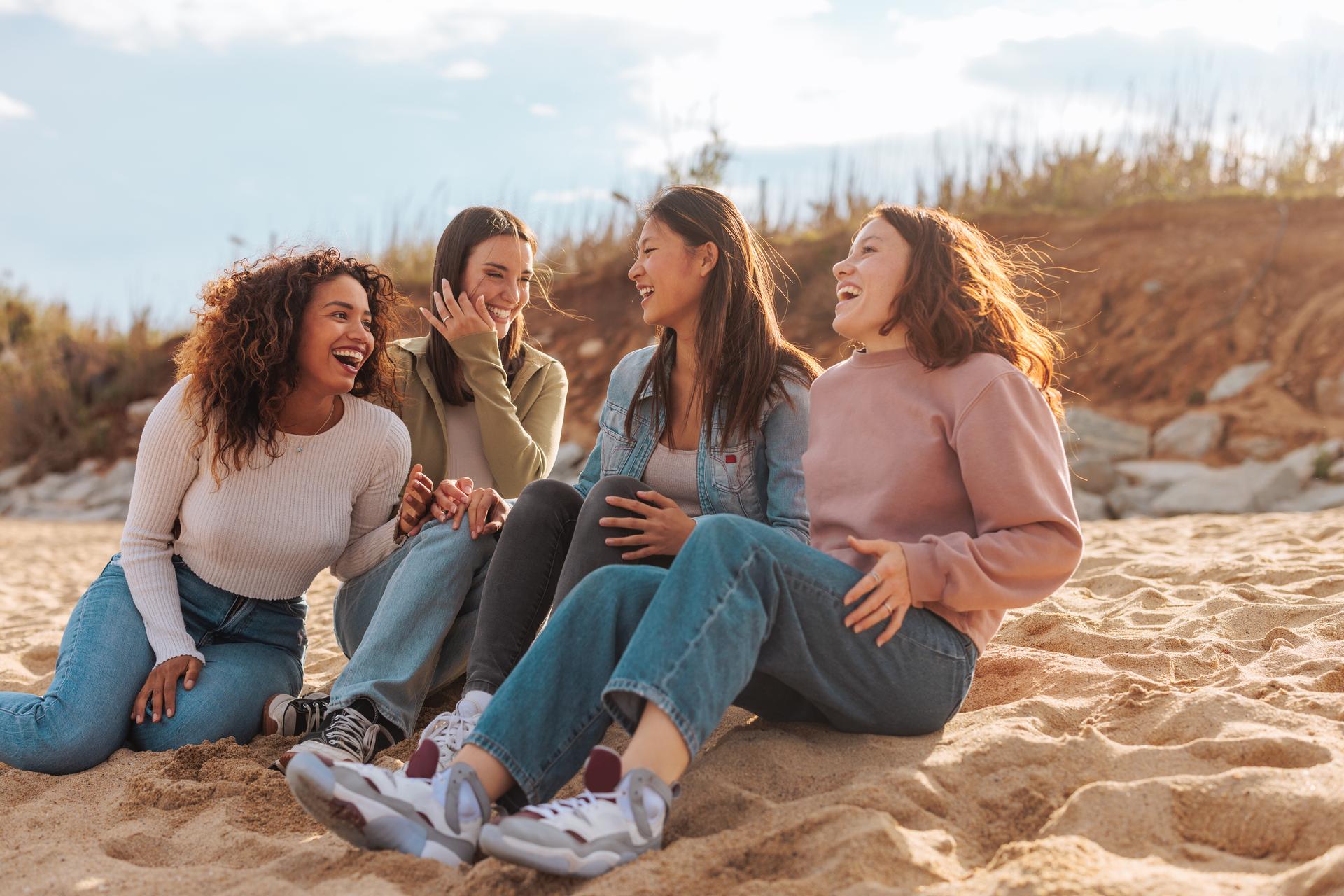 Quattro giovani donne sedute sulla sabbia che ridono insieme, simbolo di relazioni tra donne
