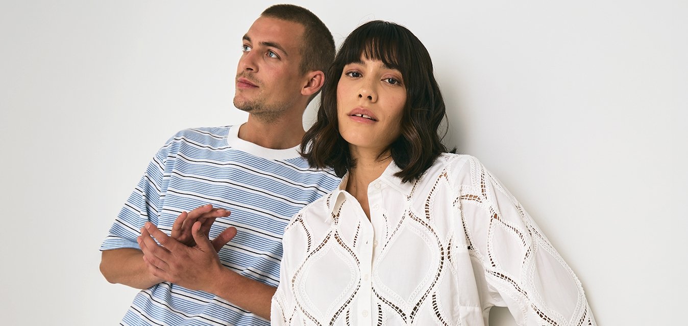 Maglietta a righe blu e bianca abbinata a pantaloni beige, insieme a una camicia di pizzo bianca con ritagli a diamante e pantaloni a righe. Sfondo puro.