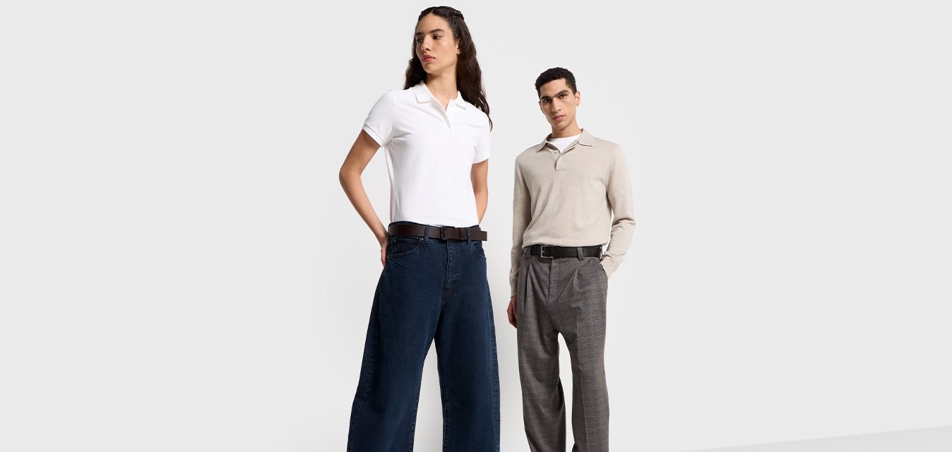 Due modelle si trovano di fronte a uno sfondo grigio chiaro. Una indossa una polo bianca e jeans larghi scuri; l'altra un maglione beige e pantaloni grigi.