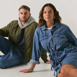 Due modelli seduti su uno sfondo neutro. Il maschio indossa una giacca bomber verde oliva, jeans blu e scarpe nere. La femmina indossa un vestito di denim con cintura annodata in vita e calzini beige con scarpe nere.