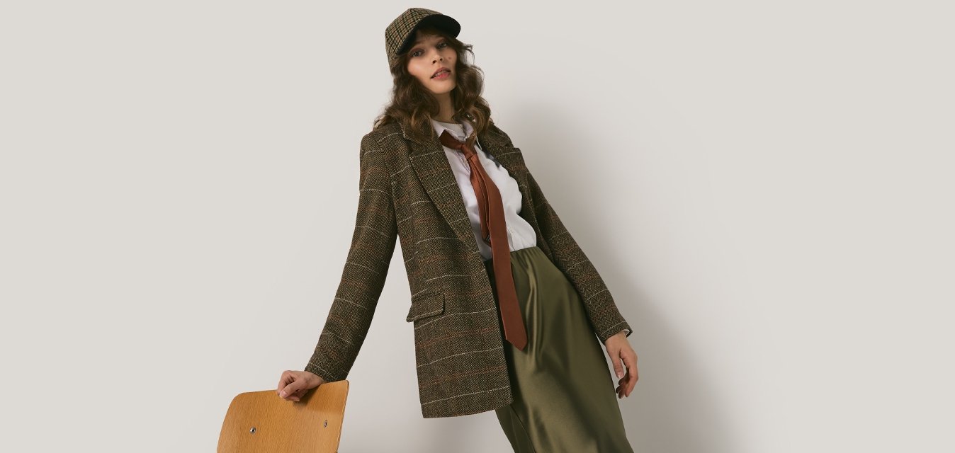 Una donna in giacca a fantasia marrone, berretto coordinato, cravatta rossa, camicia bianca e pantaloni verdi si appoggia a una sedia di legno su uno sfondo chiaro e semplice.