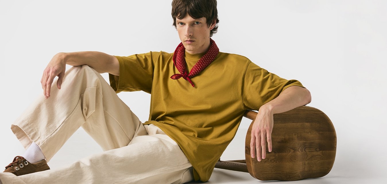 T-shirt oversize giallo senape, pantaloni beige a gamba larga, bandana rossa a pois, scarpe brown slip-on, con uno sgabello di legno rotondo.