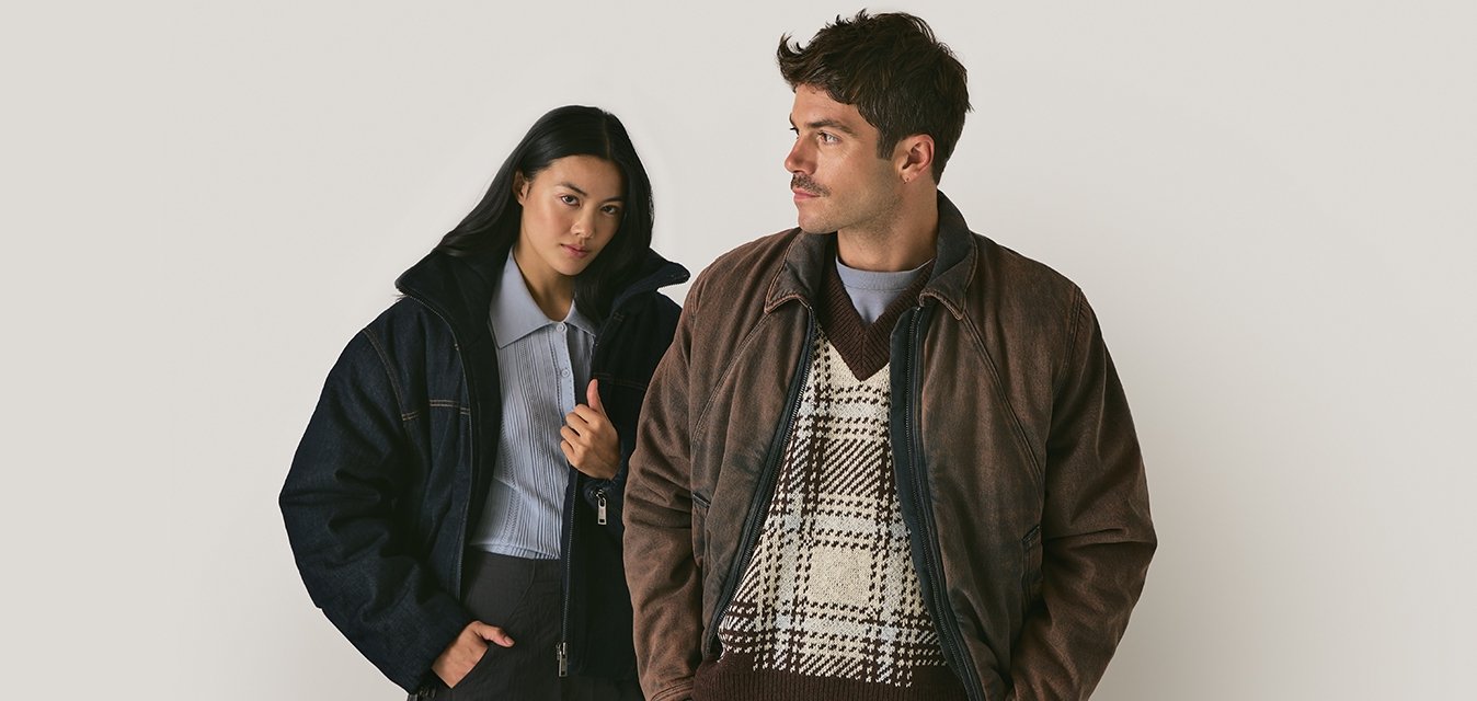 Due persone indossano giacche: una in un piumino in denim scuro e l'altra in una giacca di pelle marrone sopra un maglione a motivo. Entrambi hanno una vestibilità slim.