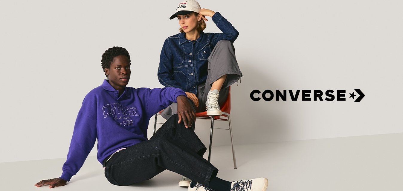 Due persone in abbigliamento casual; una seduta sul pavimento in un maglione viola, pantaloni neri e stivali bianchi; l'altra su sedia in giacca di jeans, pantaloni grigi e cappellino bianco.