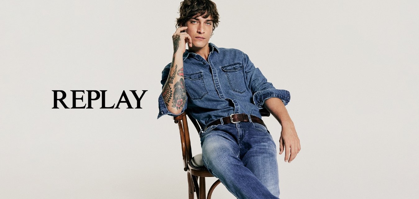 Camicia di jeans e jeans blu, indossati con una cintura marrone. Scarpe da ginnastica bianche. Seduto su una sedia di legno contro uno sfondo neutro.