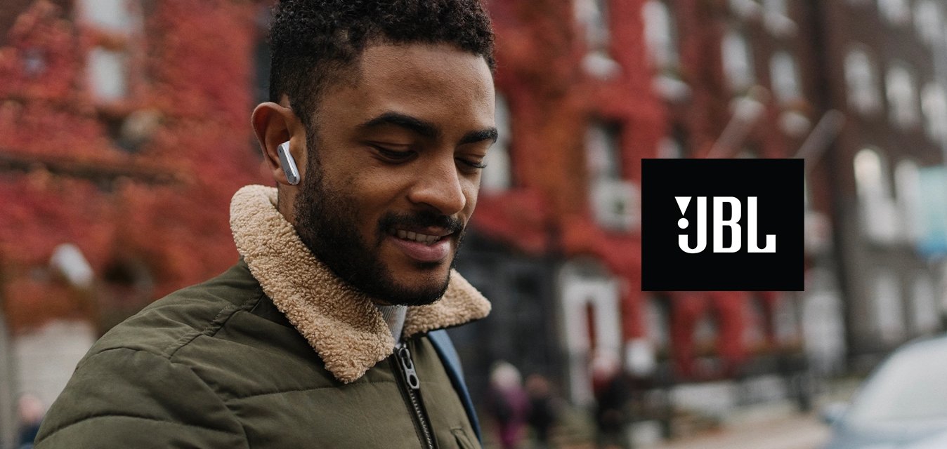 Uomo con cuffie e giacca verde guarda lo smartphone su una strada di città con edifici dalle foglie rosse e un'auto parcheggiata sullo sfondo.