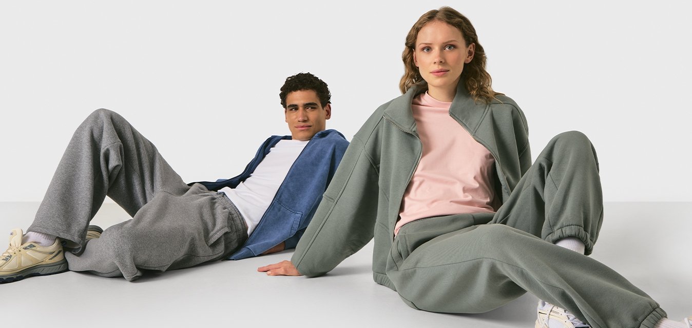 Due persone che indossano pantaloni comodi e giacche si rilassano sedute per terra contro uno sfondo chiaro e semplice.