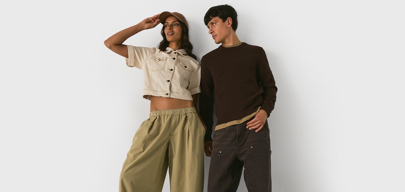 Una donna e un uomo stanno fianco a fianco contro un muro bianco; lei indossa una maglietta cropped beige e pantaloni larghi, lui indossa un maglione marrone e pantaloni scuri.