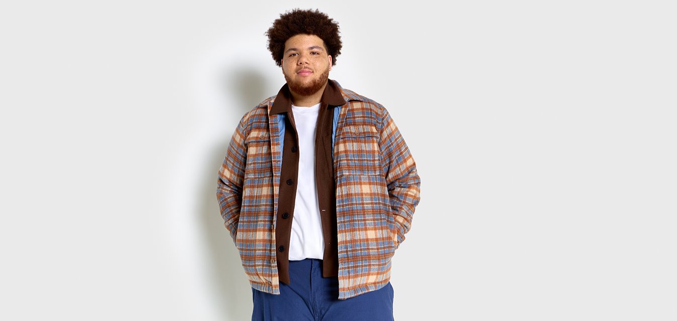 Giacca a quadri con tonalità marroni, blu e arancioni, chiusura frontale con bottoni e due tasche sul petto, indossata sopra una camicia marrone e una t-shirt bianca.