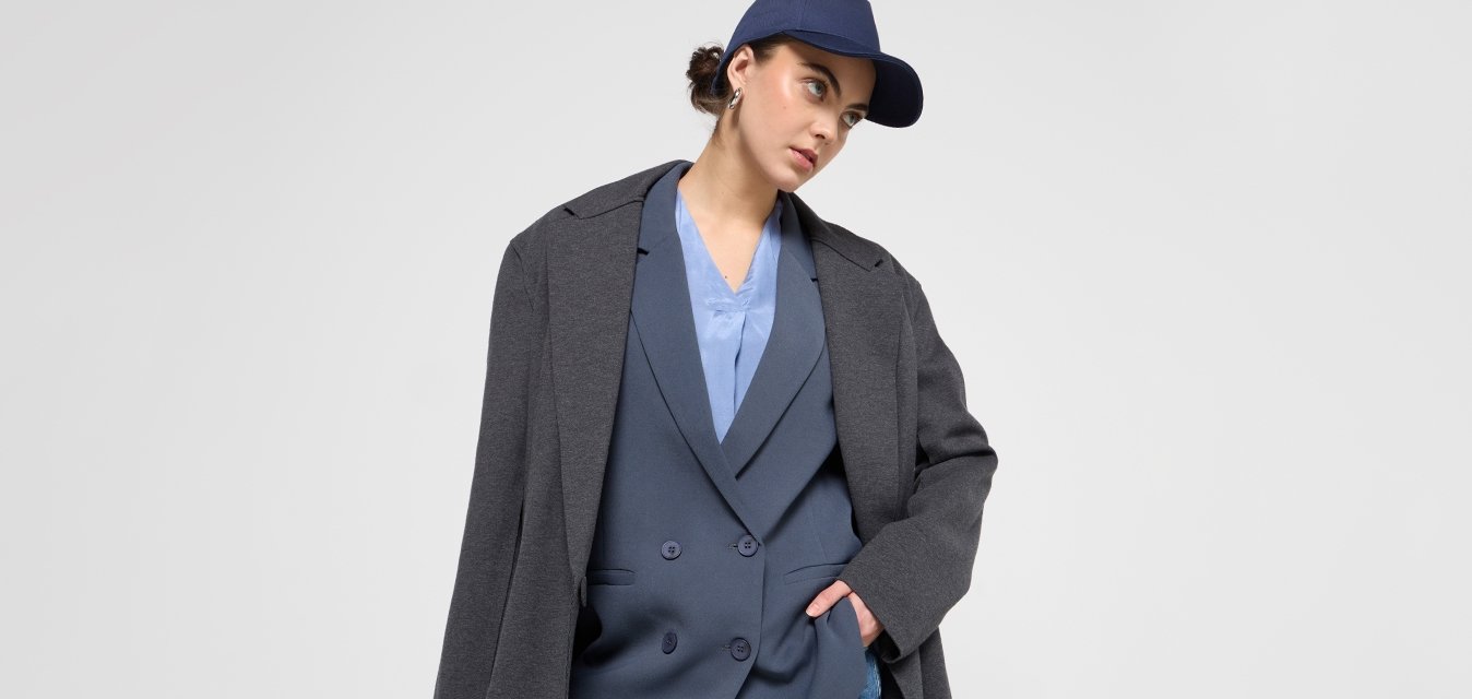 Blazer grigio oversize su un gilet blu doppiopetto, abbinato a pantaloni cargo blu. Il modello indossa un berretto blu navy, con uno sfondo a texture liscia.