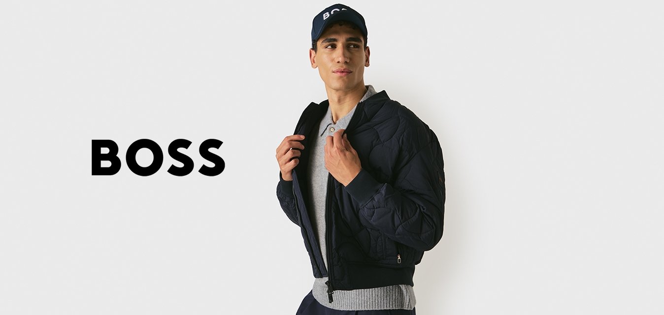 Giovane uomo che indossa un giubbotto imbottito nero, un maglione grigio, pantaloni blu navy e un cappellino blu navy, mentre sistema il colletto del giubbotto, guardando a destra, su uno sfondo chiaro e semplice.