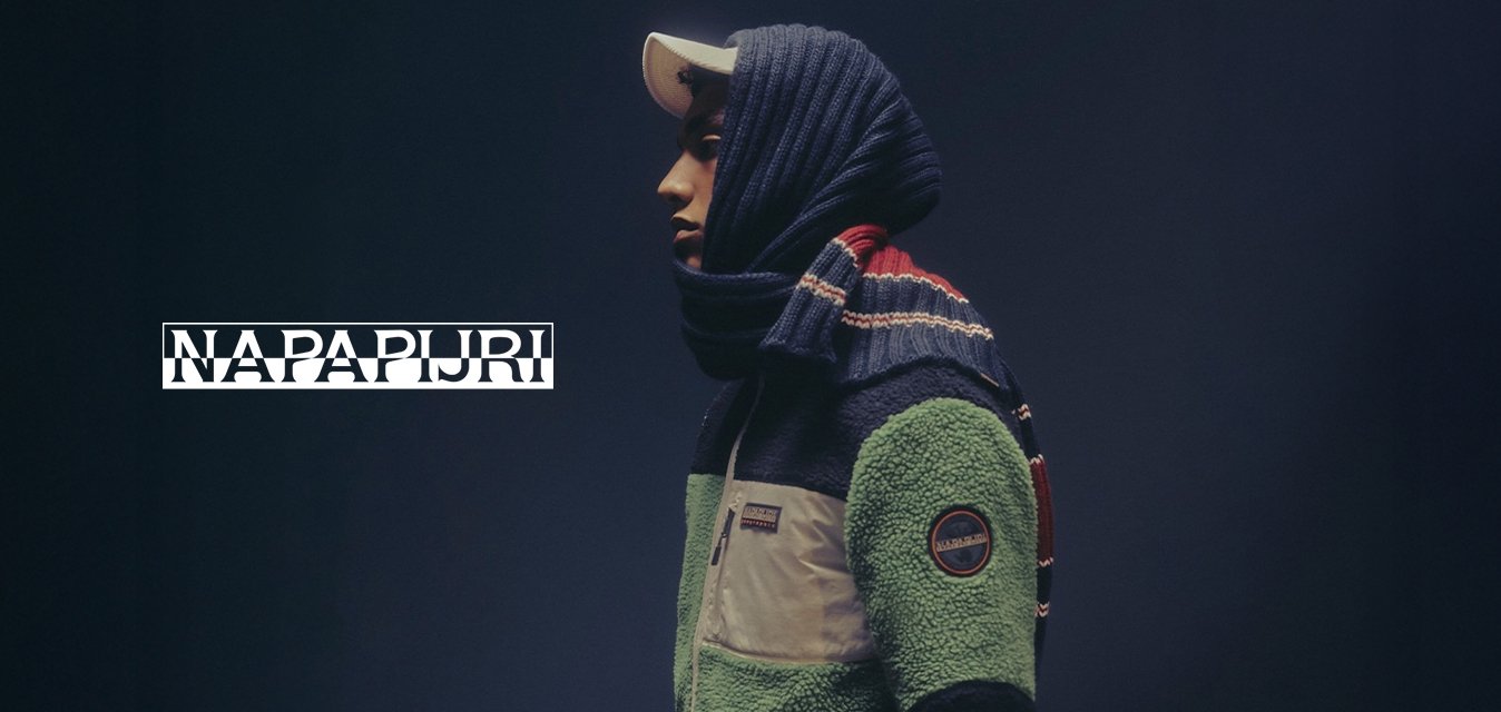 Persona di profilo che indossa una giacca di pile verde e nera, un cappello bianco e una sciarpa blu con strisce rosse e bianche su uno sfondo scuro.