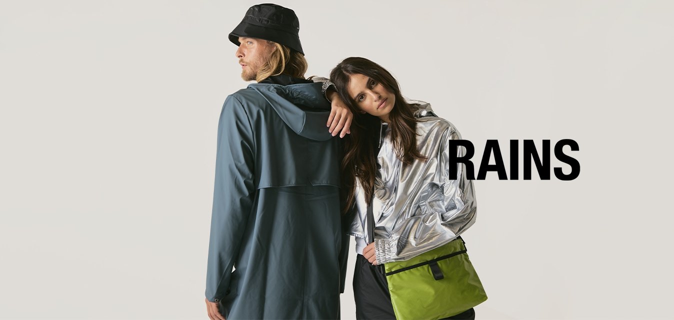 Un uomo con un impermeabile blu e un cappello a secchiello nero è in piedi schiena contro schiena con una donna in giacca argento che inclina la testa sulla sua spalla, mentre porta una borsa verde.