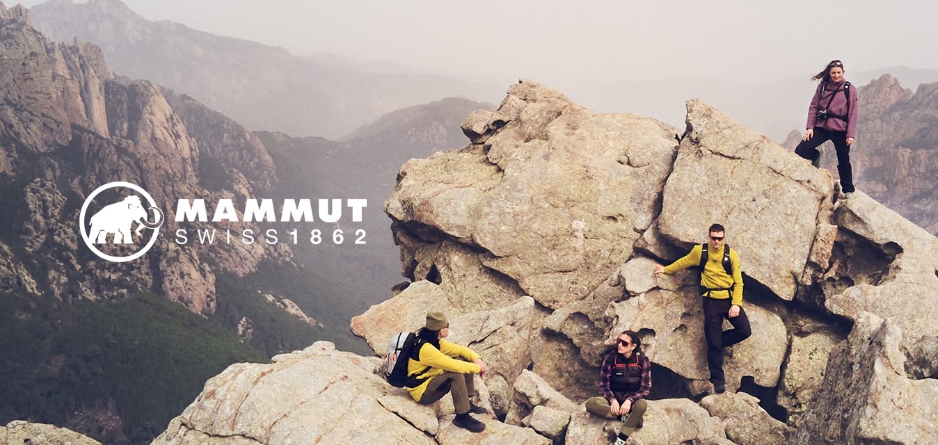 Quattro individui su un terreno roccioso, vestiti con giacche colorate. Le rocce sono textured e irregolari, con uno sfondo montuoso sotto un cielo nuvoloso.