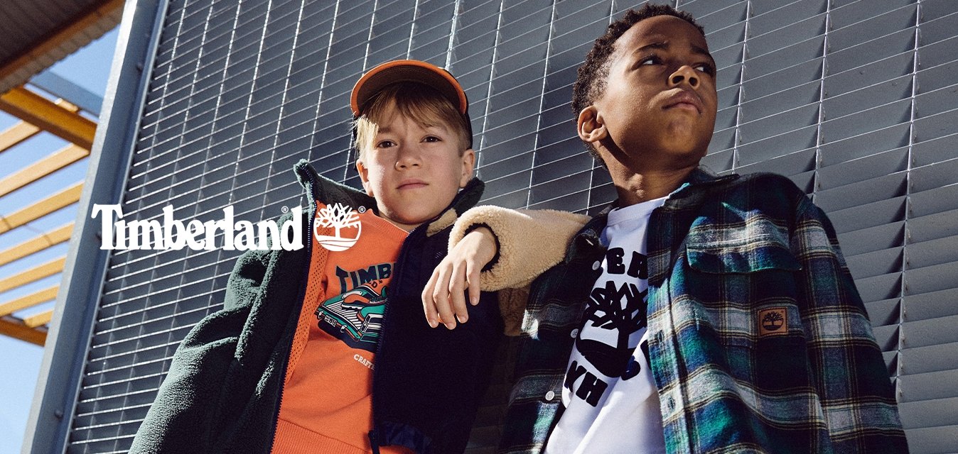 Due bambini sono in piedi contro un muro grigio con persiane. Uno indossa una giacca nera sopra una felpa arancione e jeans blu, con stivali neri. L'altro ha una giacca a quadri sopra una camicia bianca, pantaloni color khaki e scarpe gialle.