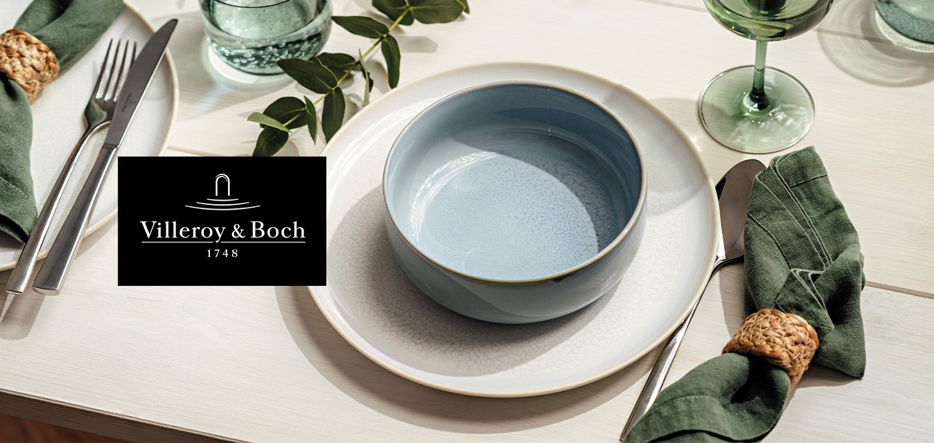 Piatti da pranzo bianchi con una ciotola blu, tovaglioli verdi in anelli intrecciati, posate, bicchieri trasparenti e verdi, e decorazione da tavolo con foglie.