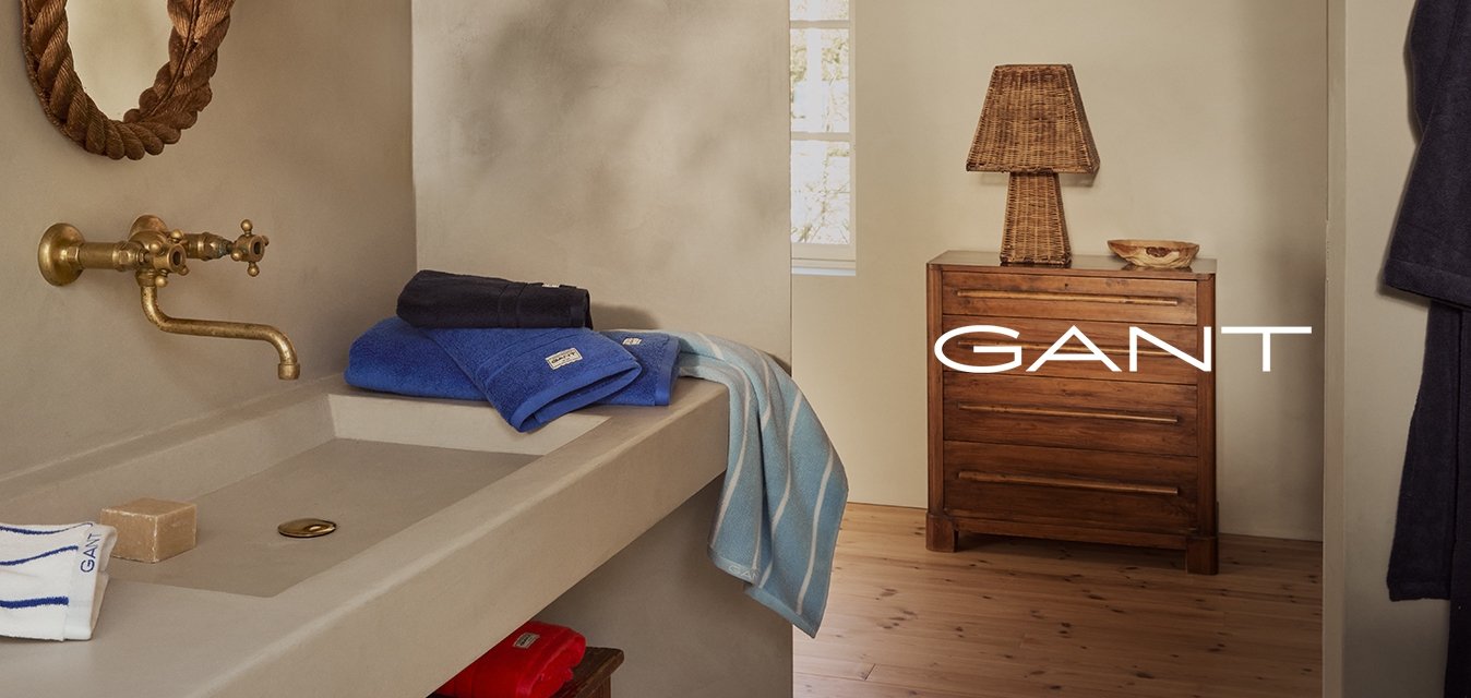 Bagno con pareti beige, uno specchio a corda curvo, un rubinetto in ottone, asciugamani impilati (blu, blu scuro e a righe), un comò in legno e una lampada intrecciata.