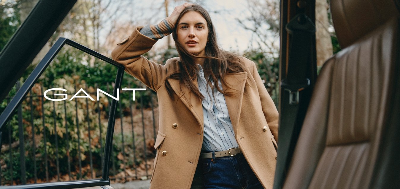Cappotto di lana tan con bottoni dorati sopra una camicia a righe blu chiaro, abbinato a jeans di denim scuri. In piedi accanto a una portiera di un'auto aperta.