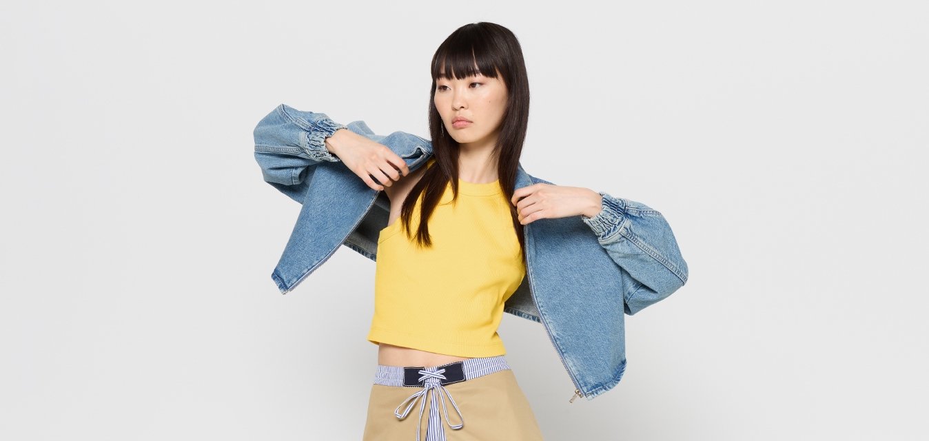 Giacca di denim con maniche a sbuffo, top giallo corto e gonna beige con una cravatta a righe. Texture morbide; contrasto di colori vivaci.