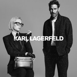 Donna con capelli biondi corti e occhiali seduta su uno sgabello, indossa un completo, guanti e mocassini mentre tiene una borsa; un uomo in piedi accanto a lei indossa un abbigliamento casual scuro.