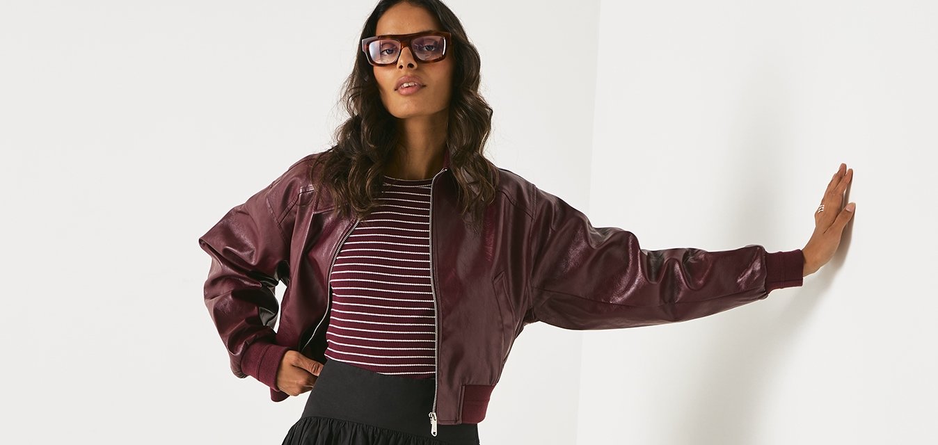 Giubbotto bomber in pelle bordeaux, maglietta a righe, gonna nera plisettata, stivali neri con plateau fino al ginocchio. La modella posa contro un muro bianco.