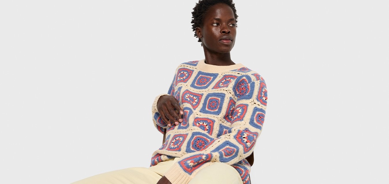 Maglione a maglia con motivi geometrici multicolori in blu, rosso e crema. Presenta un colletto rotondo e polsini a coste. Indossato con pantaloni beige.