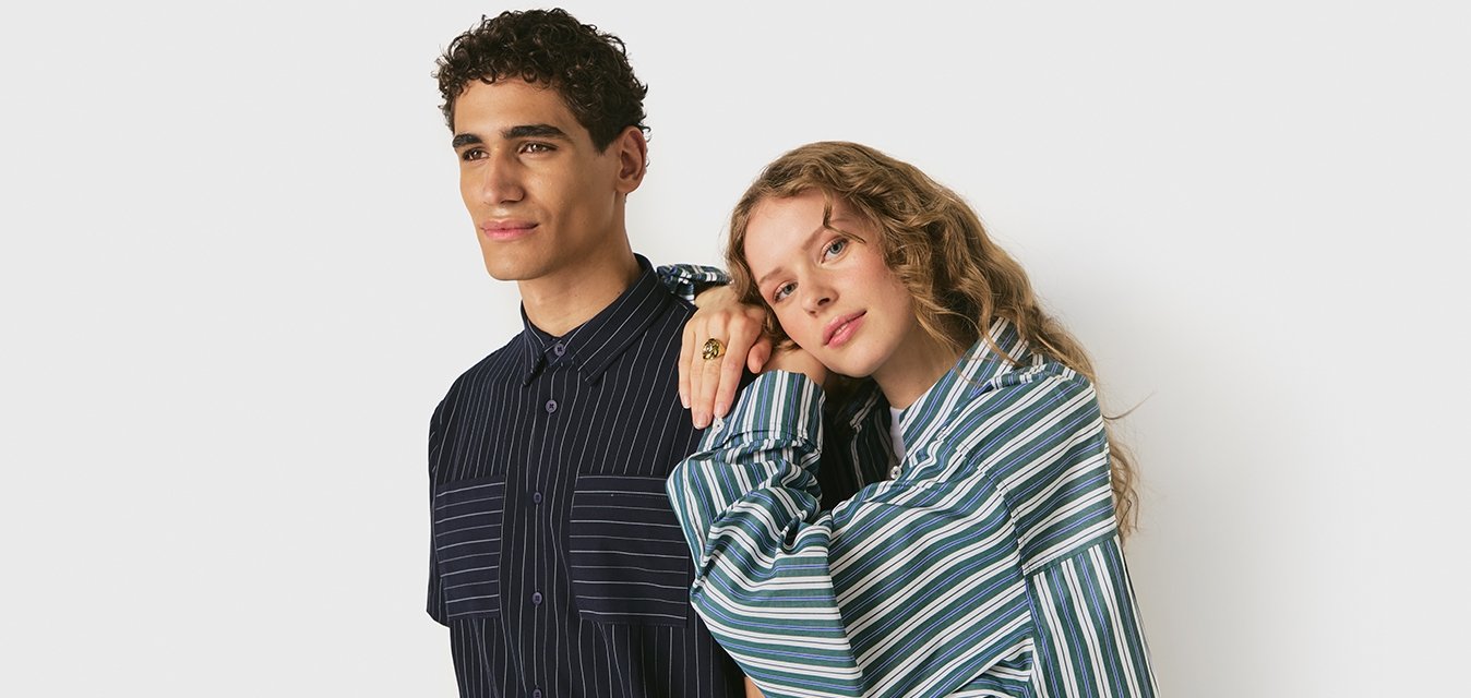 Un uomo e una donna sono in piedi uno accanto all'altro, entrambi indossano camicie a righe; la donna poggia la testa e la mano sulla spalla dell'uomo su uno sfondo bianco.