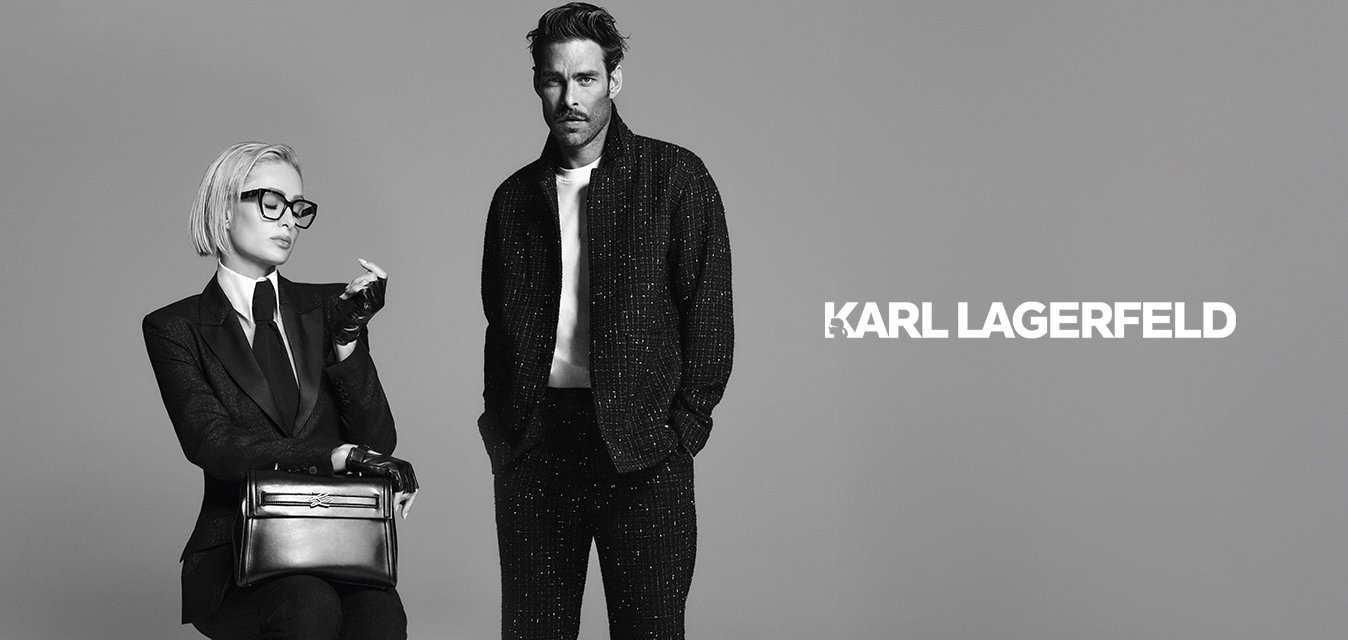 Donna con capelli biondi corti e occhiali seduta su uno sgabello, indossa un completo, guanti e mocassini mentre tiene una borsa; un uomo in piedi accanto a lei indossa un abbigliamento casual scuro.